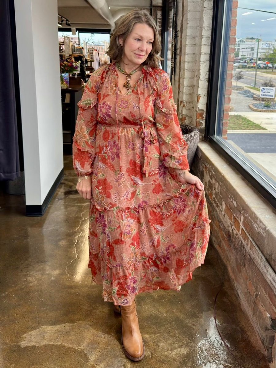 A woman wearing a long-sleeve tiered maxi dress with a red, orange, and pink floral pattern, standing in a rustic shop interior with a concrete floor and brick walls.