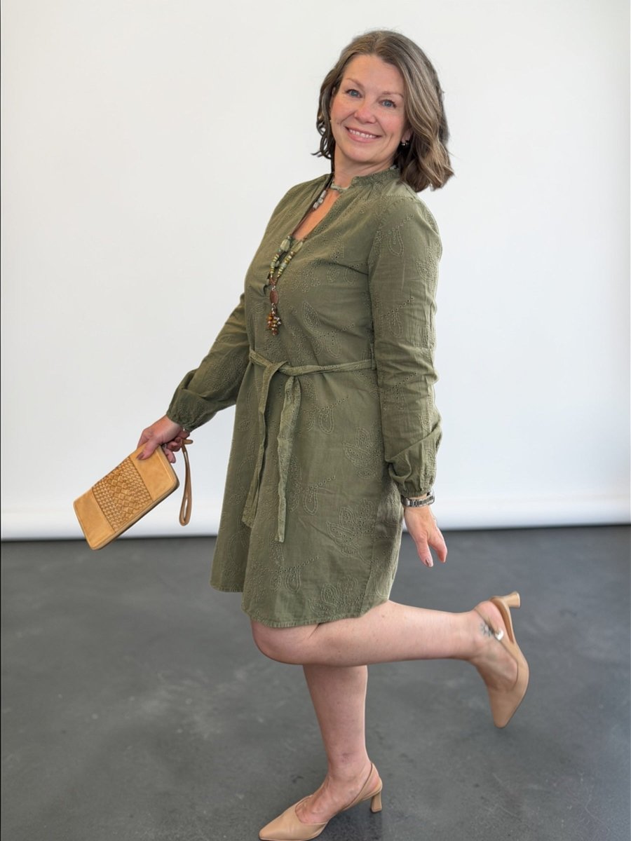 Woman in a green dress holding a beige clutch against a white wall.