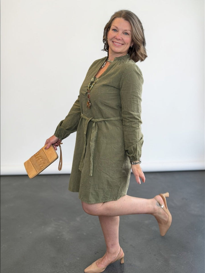 Woman in a green dress holding a beige clutch against a white wall.