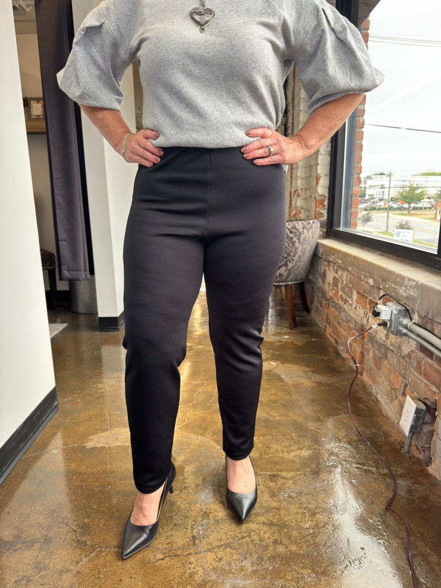 A person modeling black slim-fit pants with a gray puff-sleeve top and black pointed-toe heels, standing in a real indoor space with a brick wall, window, and polished floor.