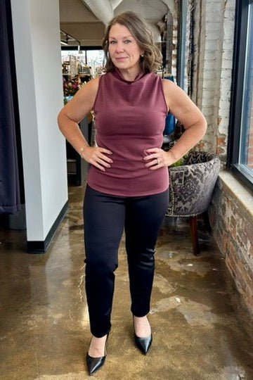 A woman modeling a sleeveless cowl-neck top in a dusty rose color, paired with black pants and black pointed-toe heels, standing in an indoor corridor with brick walls and a polished concrete floor.