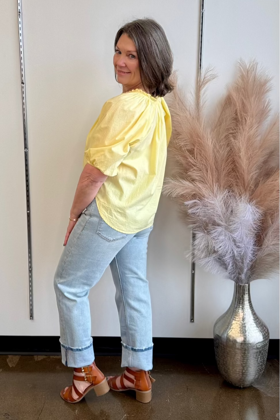 Woman looking over her shoulder wearing light wash cuffed denim and a flowy yellow shirt and a pair of brown heeled sandals.