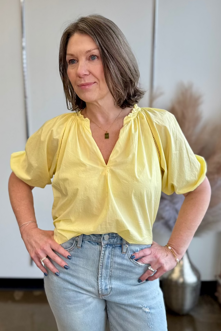 Woman wearing a pale yellow flowy top standing with her hands on hips inside looking in the distance.