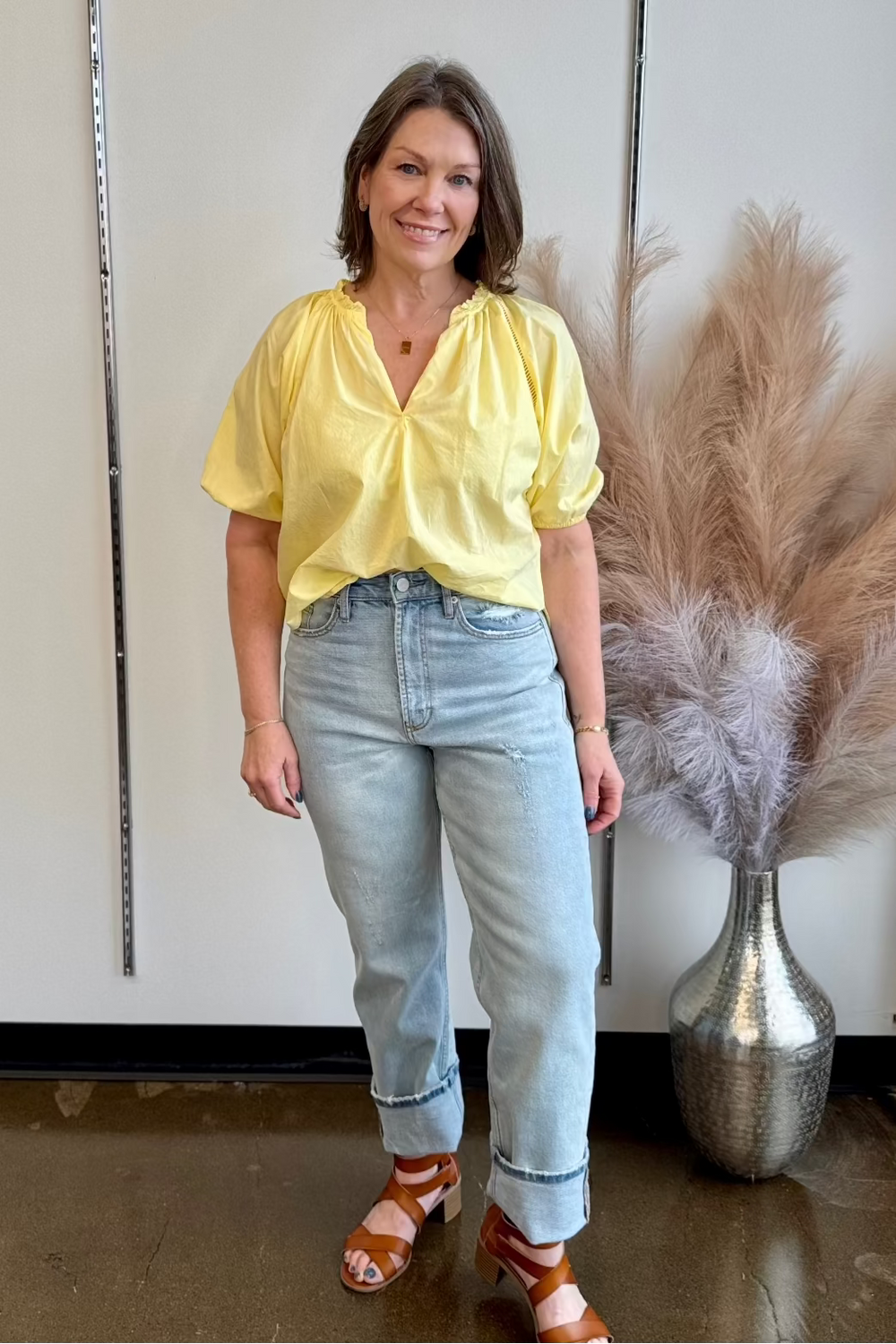Woman wearing light wash denim and a pale yellow v-neck blouse and brown sandals smiling at the camera.