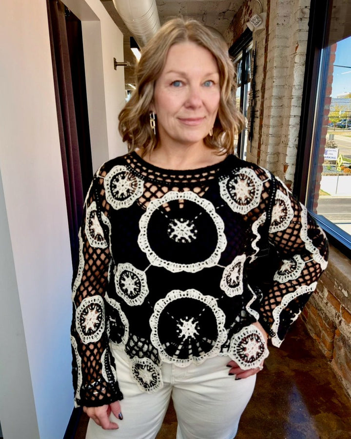 A woman wearing a black and white crocheted long-sleeve top with white pants, standing in an indoor hallway with brick walls and a window.