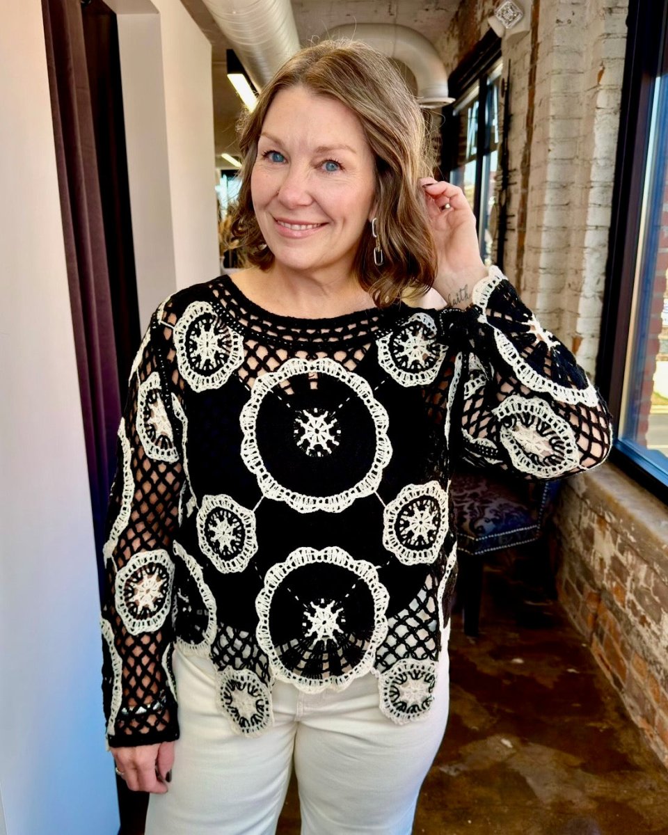 A woman wearing a black and white crochet patterned long-sleeve top with white pants, standing in an indoor setting with brick walls and windows.