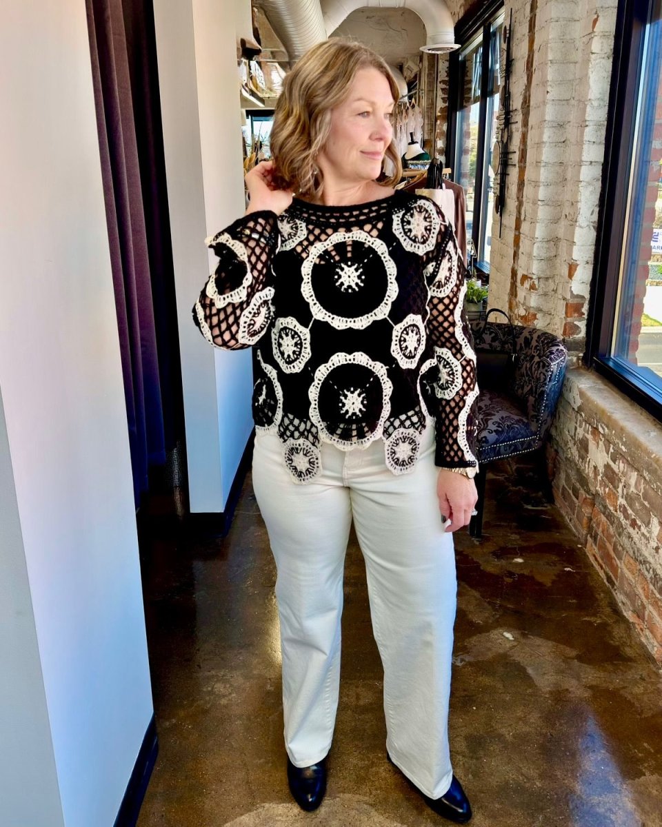 A woman modeling a black crochet knit sweater with white circular patterns, paired with off-white pants and black shoes, standing in a hallway with brick walls and windows.