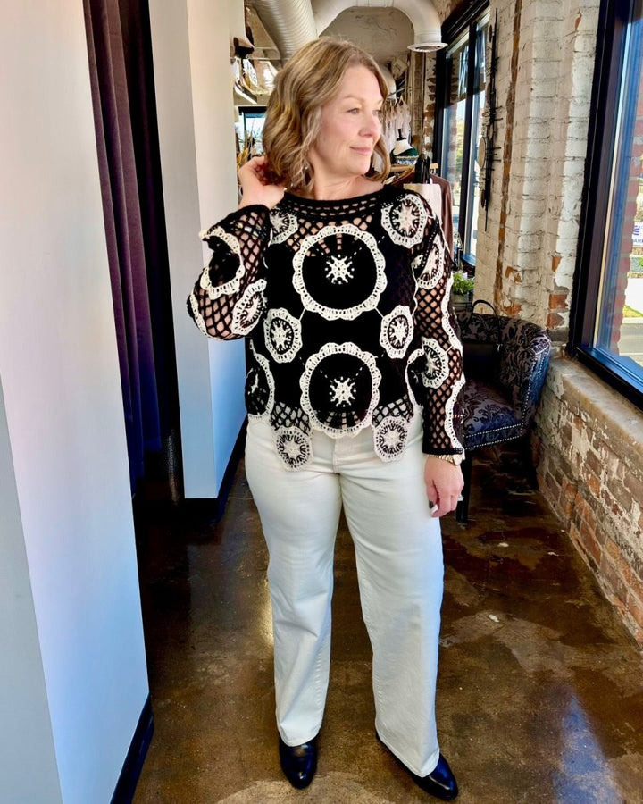 A woman modeling a black crochet knit sweater with white circular patterns, paired with off-white pants and black shoes, standing in a hallway with brick walls and windows.