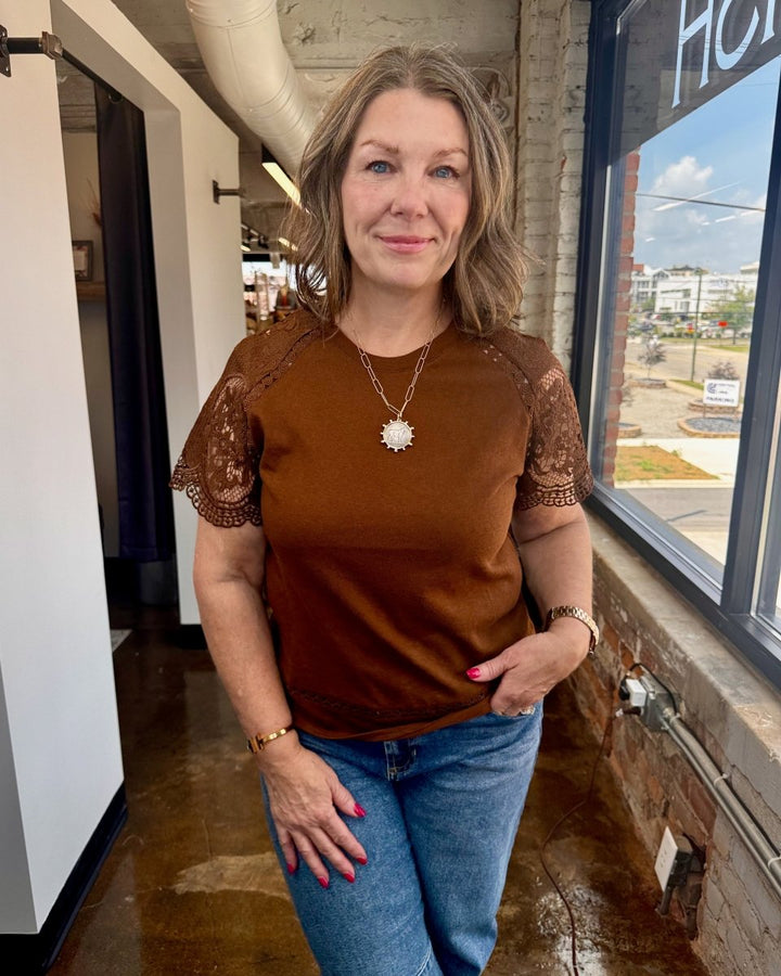 Woman wearing a brown lace-trimmed top and blue jeans standing indoors.