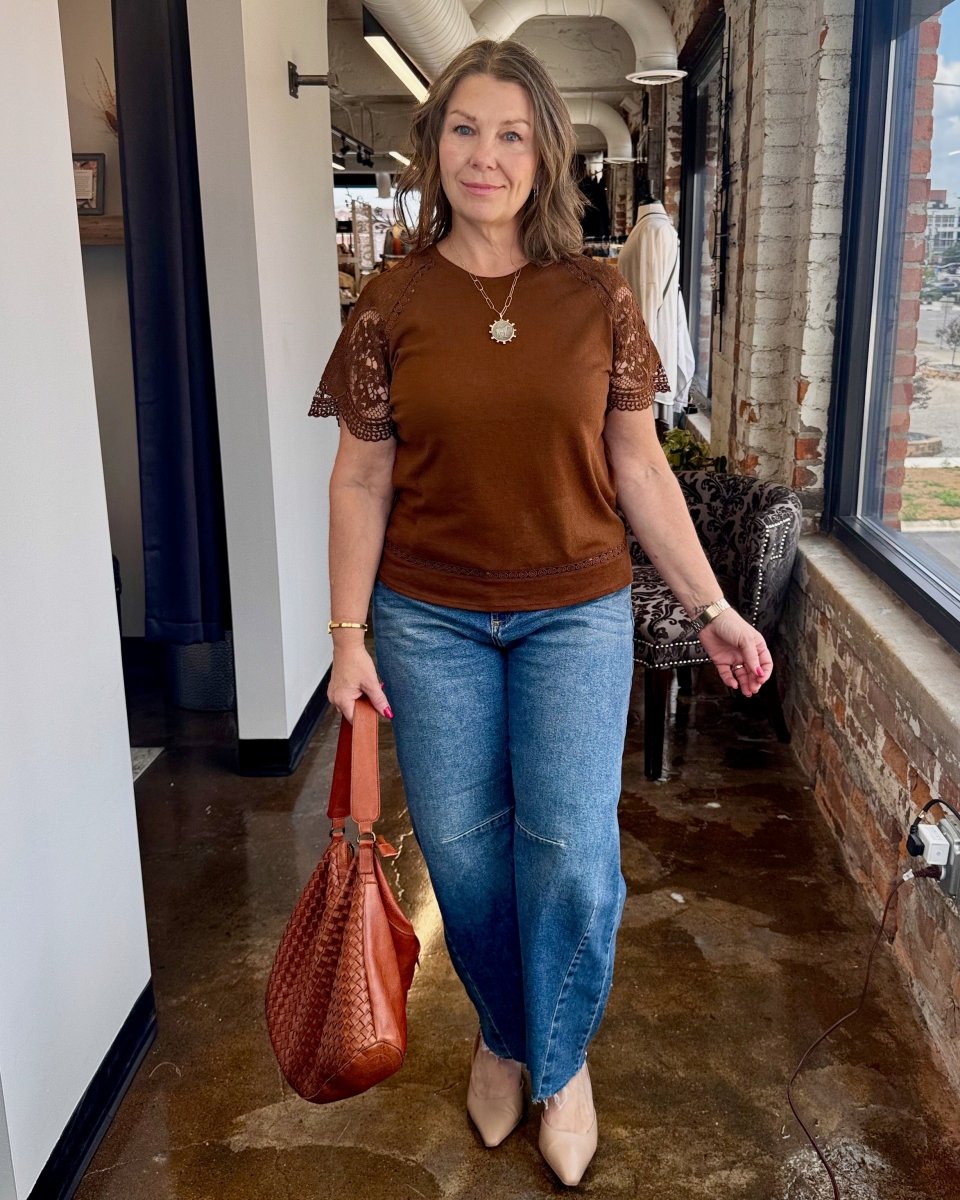 Woman in brown top and blue jeans holding a brown bag indoors.