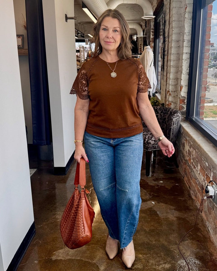 Woman in brown top and blue jeans holding a brown bag indoors.