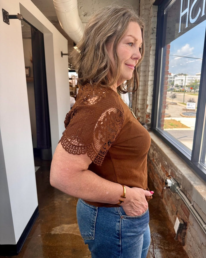 Woman wearing a brown lace top and blue jeans standing indoors.