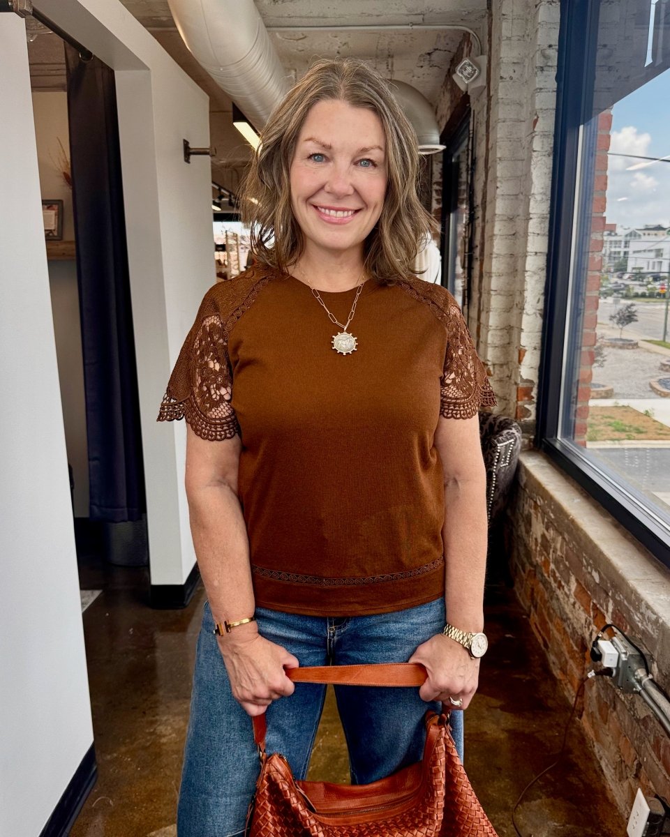 Woman wearing a brown lace-top shirt and blue jeans, holding a brown leather bag indoors.