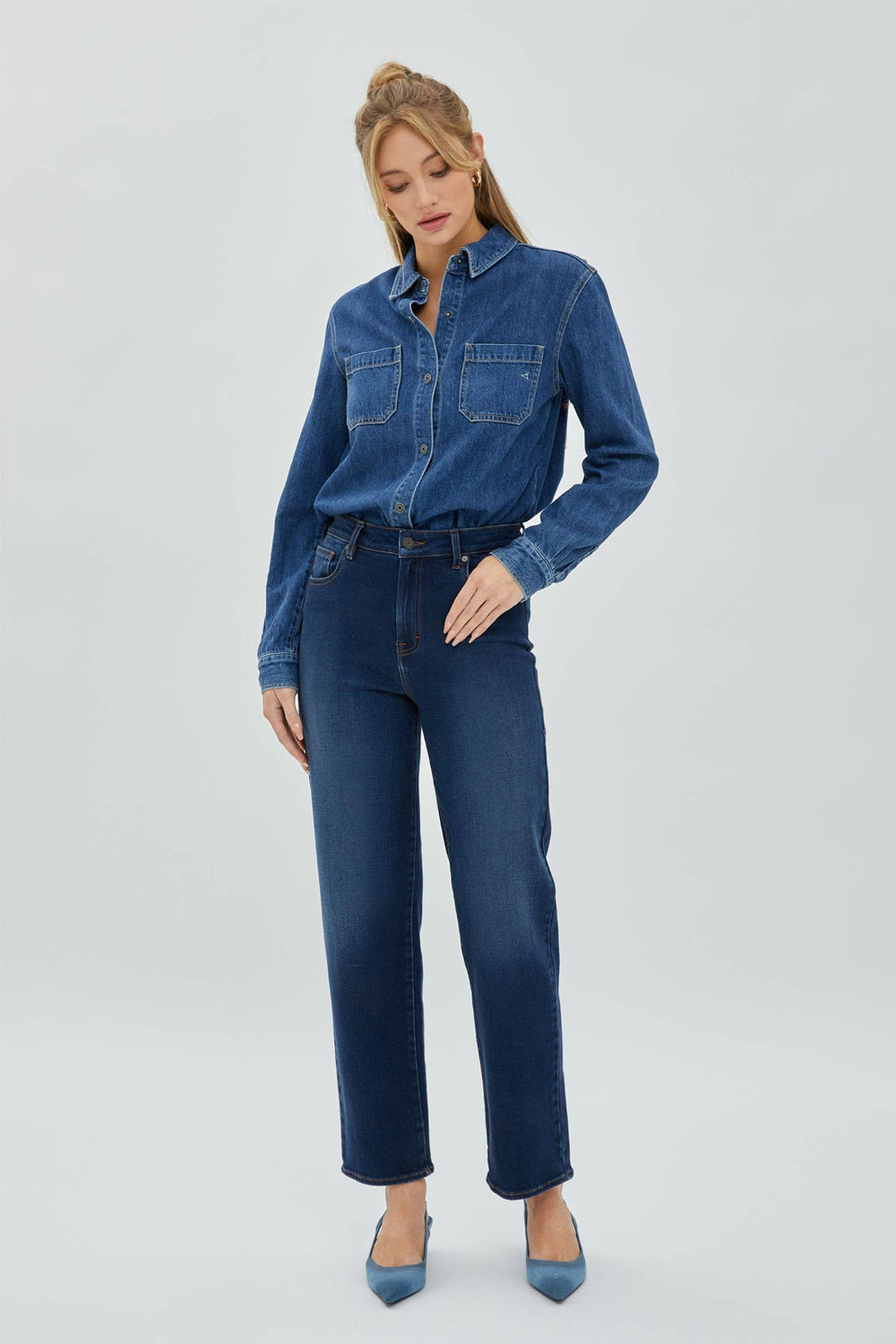 A person standing wearing straight-leg blue jeans and a denim shirt tied at the waist.