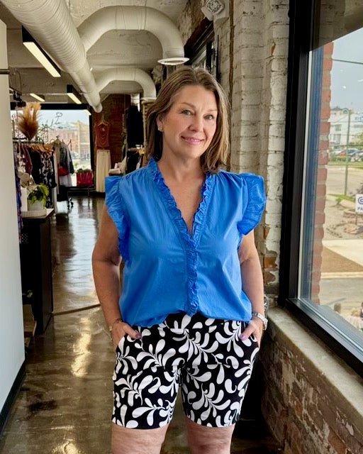 Our model is wearing a size Small in the azure blue Ellie Sleeveless Ruffle Button-Up Top by Dear John Denim, styled full-length with black and white printed bermuda shorts, hands in pockets, in a boutique setting.