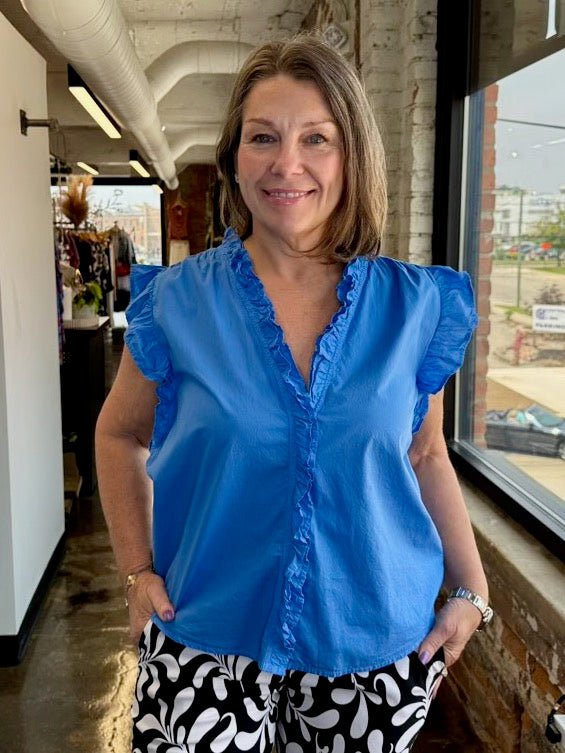 Our model is wearing a size Small in the azure blue Ellie Sleeveless Ruffle Button-Up Top by Dear John Denim, shown from the waist up with flutter ruffle sleeves and ruffled V-neckline, paired with black and white printed shorts.