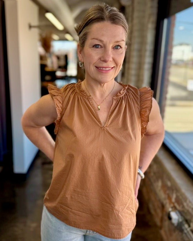 Our model is wearing a size Small in the biscuit Puja Top by Dear John Denim, shown in a close front view highlighting the V-neck, smocked ruffle neckline, and flutter sleeves, styled with a delicate gold necklace and drop earrings.