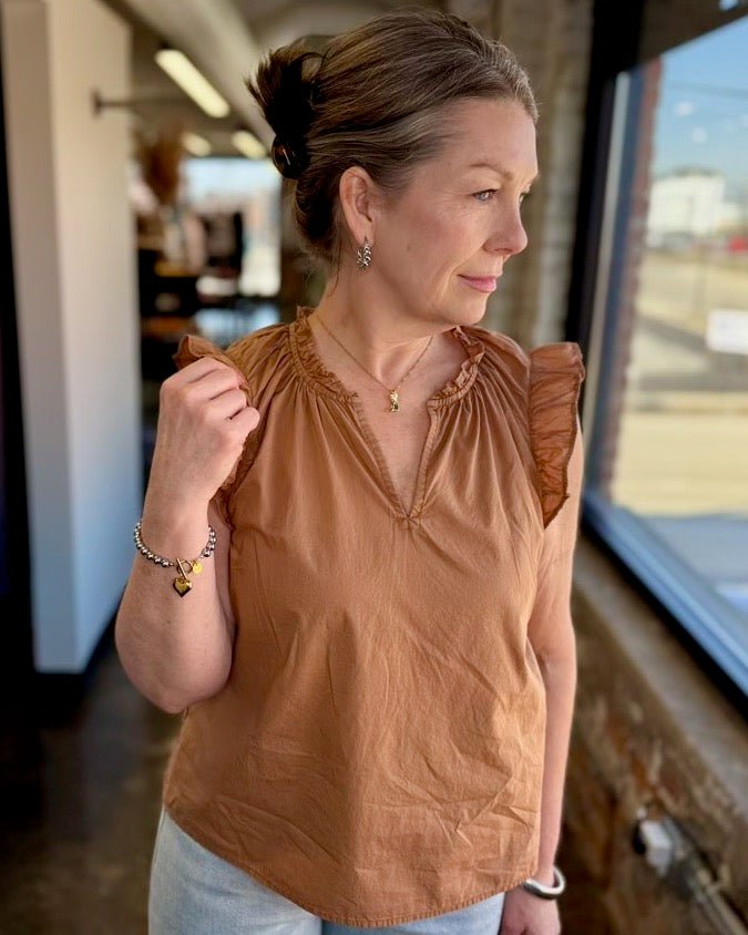 Our model is wearing a size Small in the biscuit Puja Top by Dear John Denim, shown in a three-quarter side view highlighting the flutter sleeve and smocked shoulder detail, styled with a delicate gold necklace and chain bracelet.
