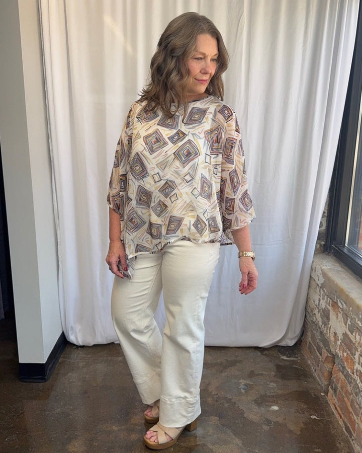 Lior Geometric Diamond Print Asymmetrical Blouse in cream with multicolor geometric diamond print, styled with white wide-leg denim and wood block heel sandals, model looking down, full length front view
