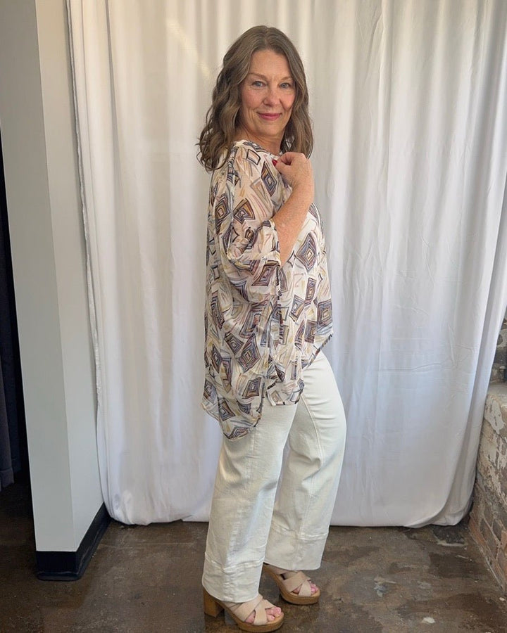 Lior Geometric Diamond Print Asymmetrical Blouse in cream with multicolor diamond print, asymmetrical drape and 3/4 sleeve detail, side view on model with white wide-leg denim
