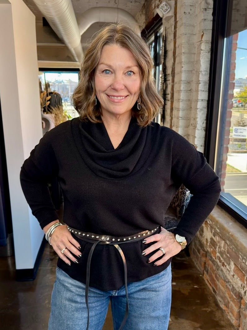 A woman with shoulder-length blonde hair stands indoors wearing a black cowl-neck knit sweater, blue jeans, and a black studded belt. She is posed with her hands on her hips in a loft-style room with brick walls and large windows.