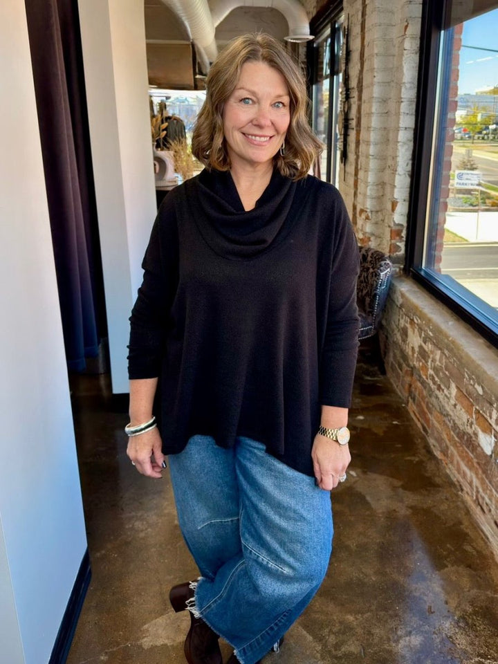 A woman wearing a black cowl-neck long-sleeve top and blue jeans standing in a shop corridor with brick walls and large windows.