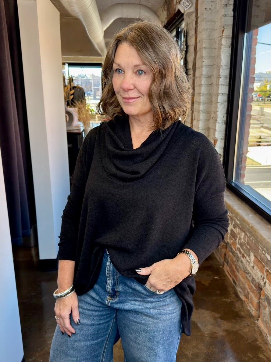 A woman with shoulder-length brown hair wearing a black cowl-neck long-sleeve top and blue jeans, standing in a loft-style room with a brick column and large windows.