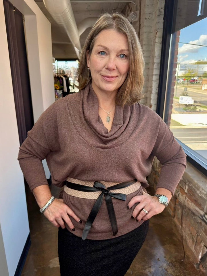 A woman wearing a brown cowl-neck knit sweater with a beige waistband and black ribbon belt, paired with a black skirt, standing in a store interior.