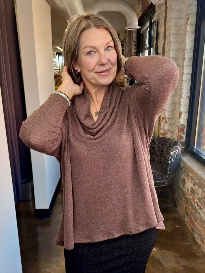 A woman wearing a dusty rose-brown cowl-neck long-sleeve top, posing in a loft-style room with brick walls and windows.