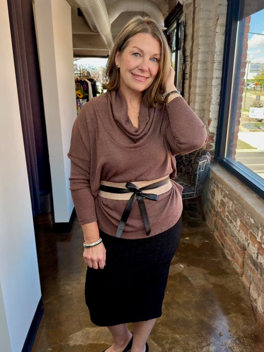 A woman modeling a brown cowl neck long-sleeve top with a beige and black striped belt, paired with a black skirt, standing in a store corridor.