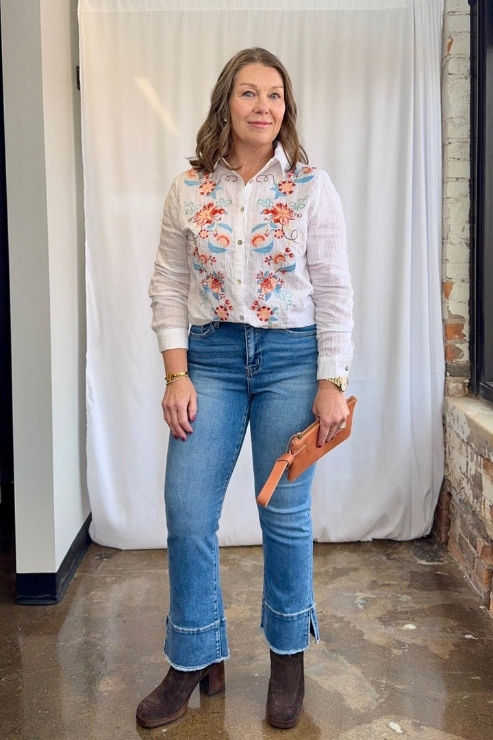 Woman wearing a floral blouse, jeans, and brown boots standing against a white curtain.