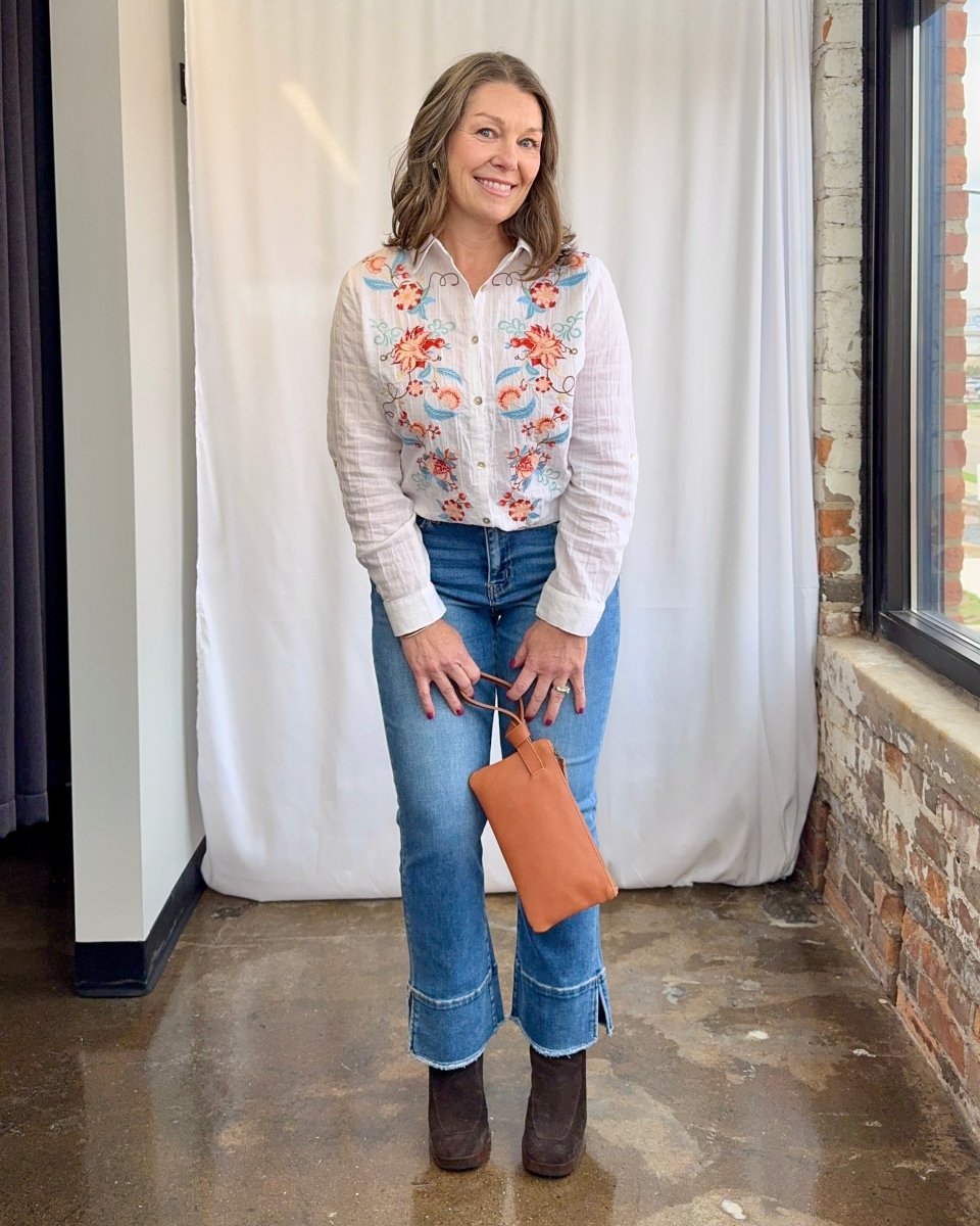 Woman wearing a floral shirt, jeans, and brown boots holding a tan clutch.