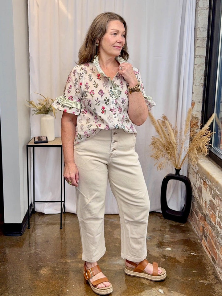 White floral block print top with green striped ruffle collar and flutter sleeves, paired with cream barrel-leg jeans and tan platform sandals at Modish Maven Boutique