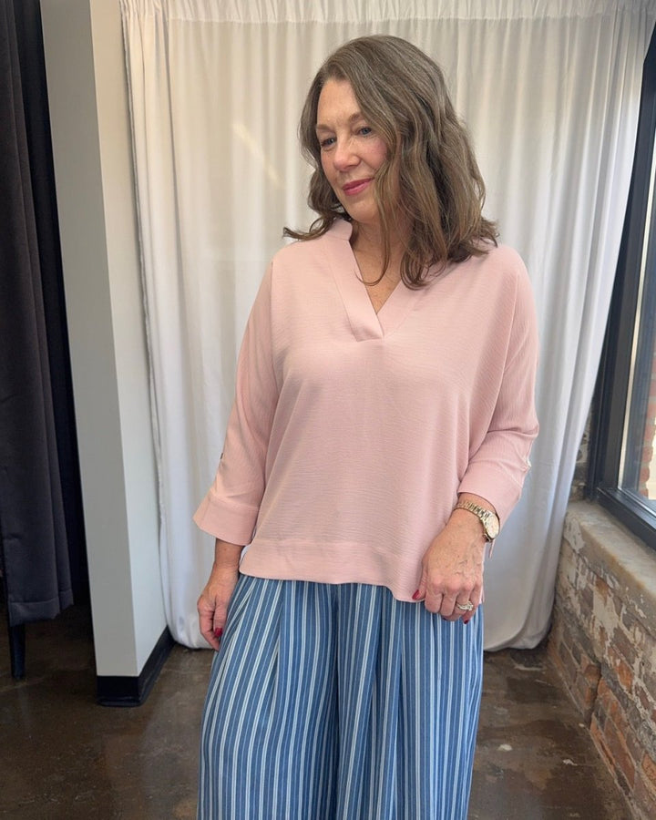Frida Crossover V-Neck Tunic Blouse in blush pink, relaxed 3/4 sleeve with button cuff detail and split hem, styled with blue stripe wide-leg trousers, close front view on model
