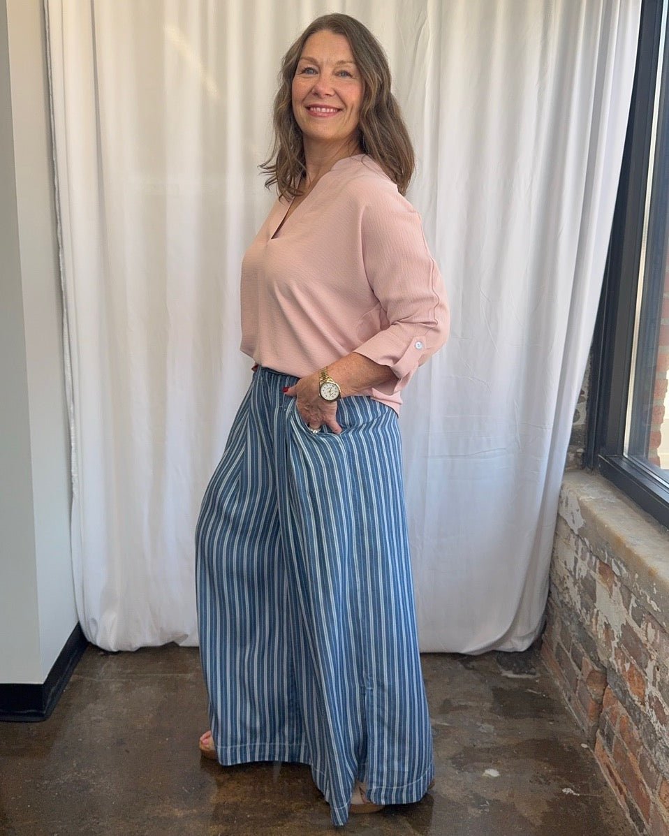 Frida Crossover V-Neck Tunic Blouse in blush pink with relaxed drape and split hem, styled with blue stripe wide-leg trousers, smiling side view on model with hand in pocket

