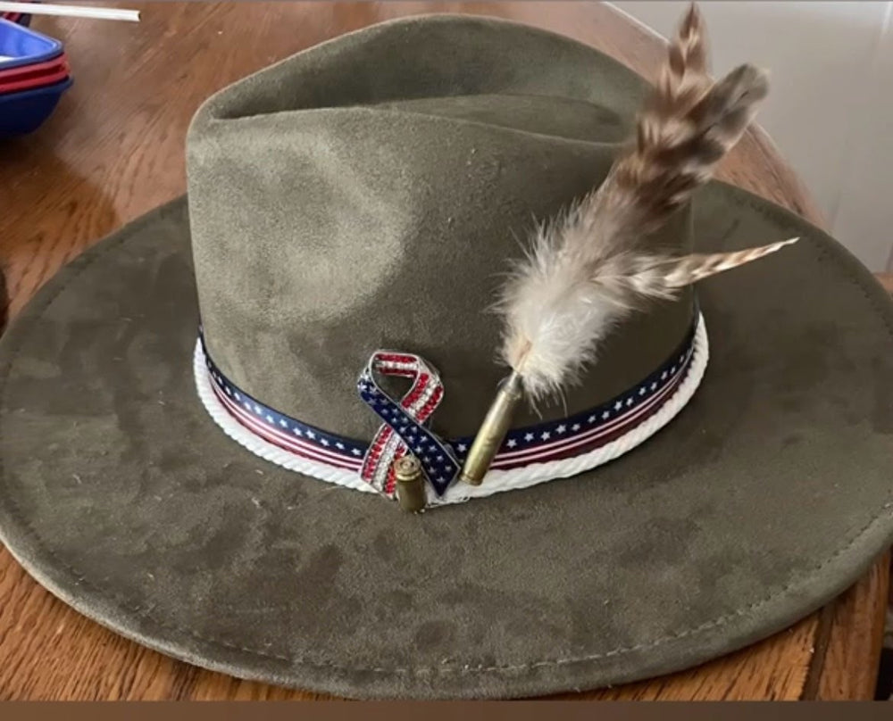 An olive green felt wide-brim hat with a patriotic red, white, and blue ribbon band and a brown feather, shown on a wooden table.
