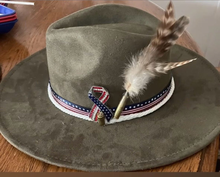 An olive green felt wide-brim hat with a patriotic red, white, and blue ribbon band and a brown feather, shown on a wooden table.