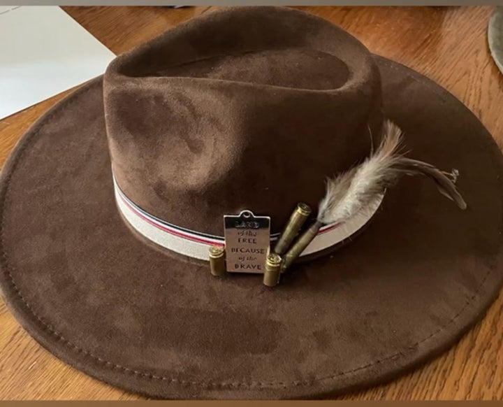 A brown suede cowboy hat with a white, red, and blue band, brass bullet accents, and a feather, displayed on a wooden table.