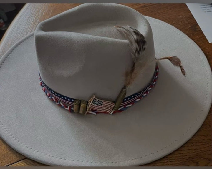 A light beige wide-brim fedora hat with a decorative band featuring a small American flag, rifle-shaped buckles, and a feather accent, resting on a wooden surface.