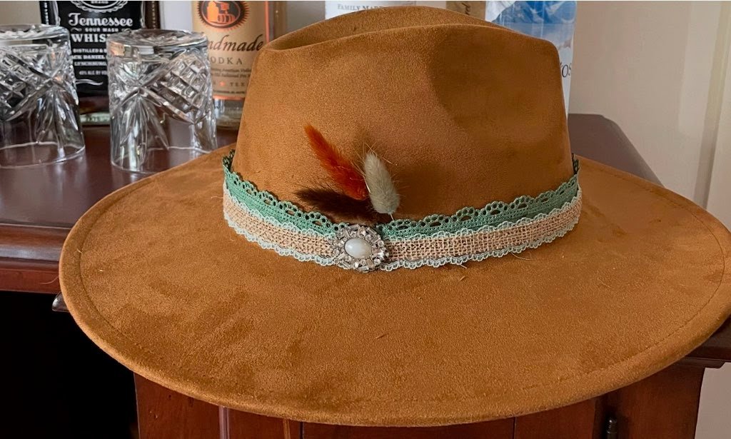A brown felt wide-brim hat with a decorative band featuring lace, a pearl-like center, and two feathers, displayed on a wooden surface with glasses and bottles in the background.