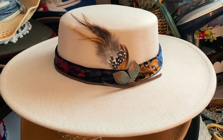 A cream-colored felt wide-brim hat with a dark patterned band and a feather accent, displayed on a stand.