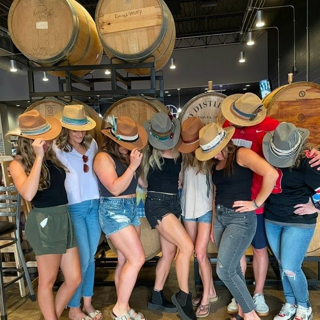 A group of nine women wearing cowboy hats pose together in a wine cellar with wooden barrels in the background.