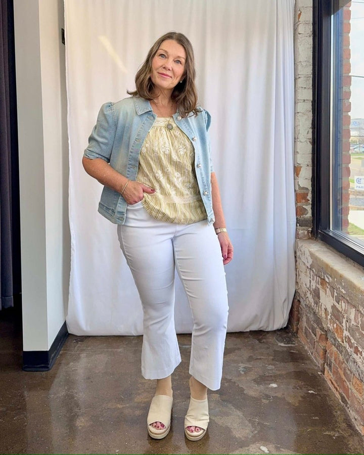 Woman wearing a denim jacket, gold top, and white pants standing indoors.