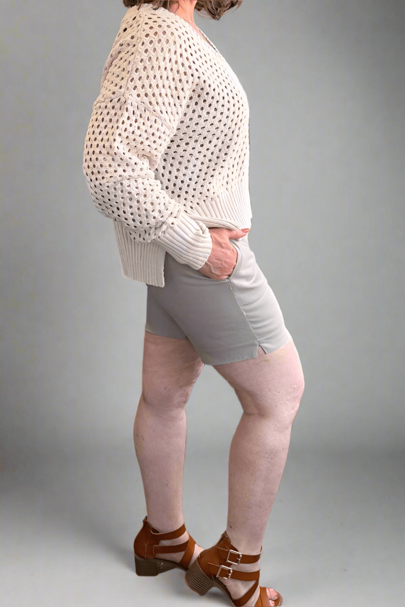 Side view of a woman wearing 'Summer Straw' high-rise stretch Millennium shorts with a 7-inch inseam, styled with a cream v-neck open-knit sweater and tan strappy sandals.
