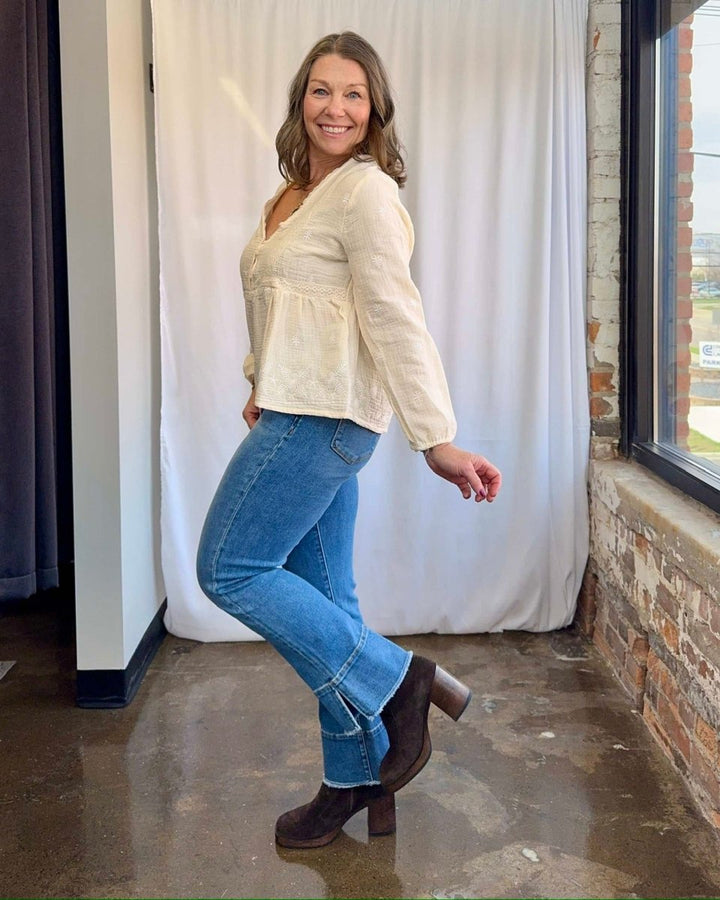 Woman in a cream blouse, blue jeans, and brown boots standing indoors.