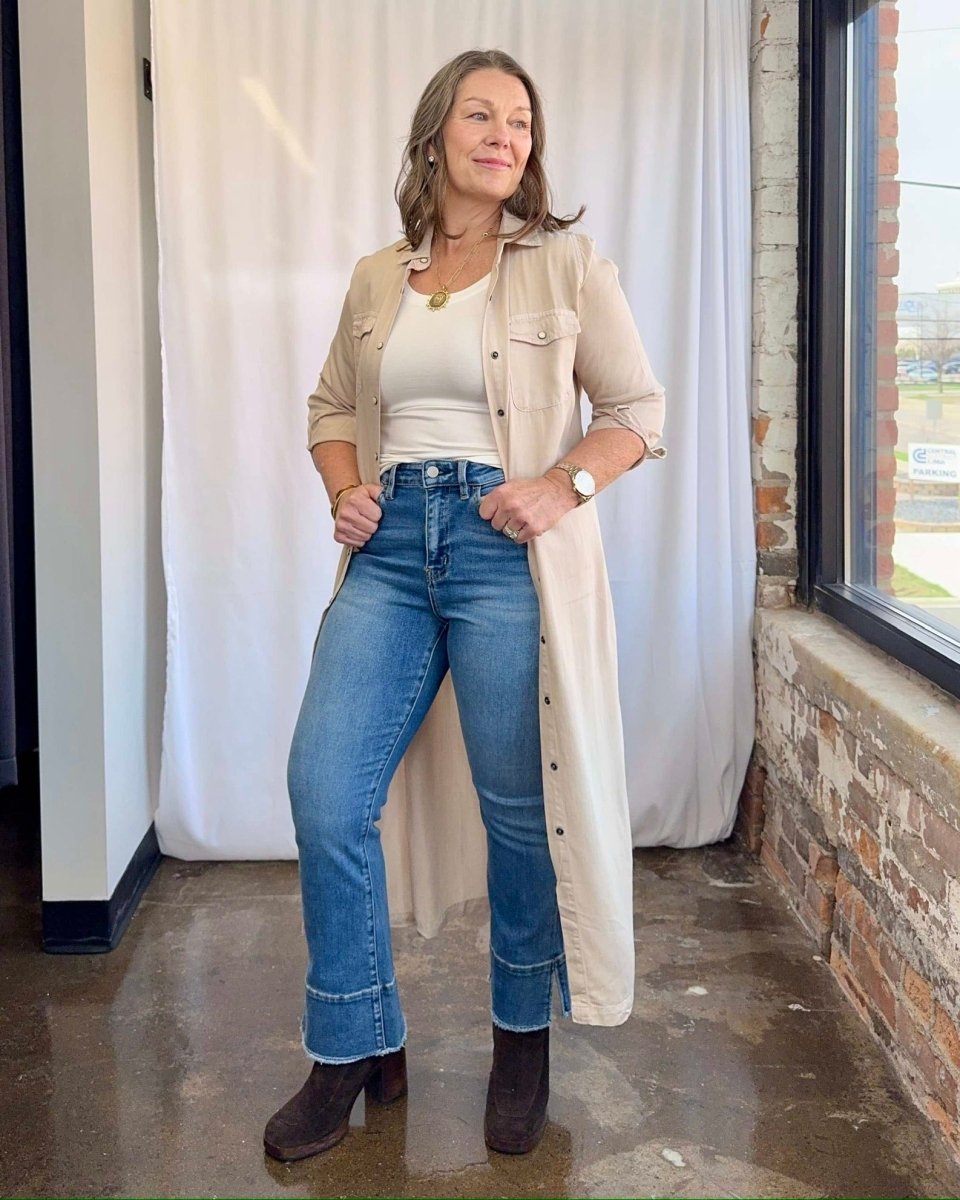 Woman wearing a beige trench coat, white top, blue jeans, and brown boots indoors.