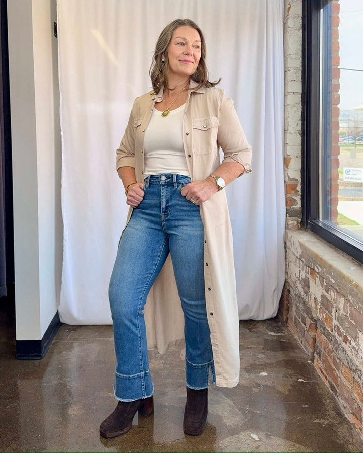 Woman wearing a beige trench coat, white top, blue jeans, and brown boots indoors.