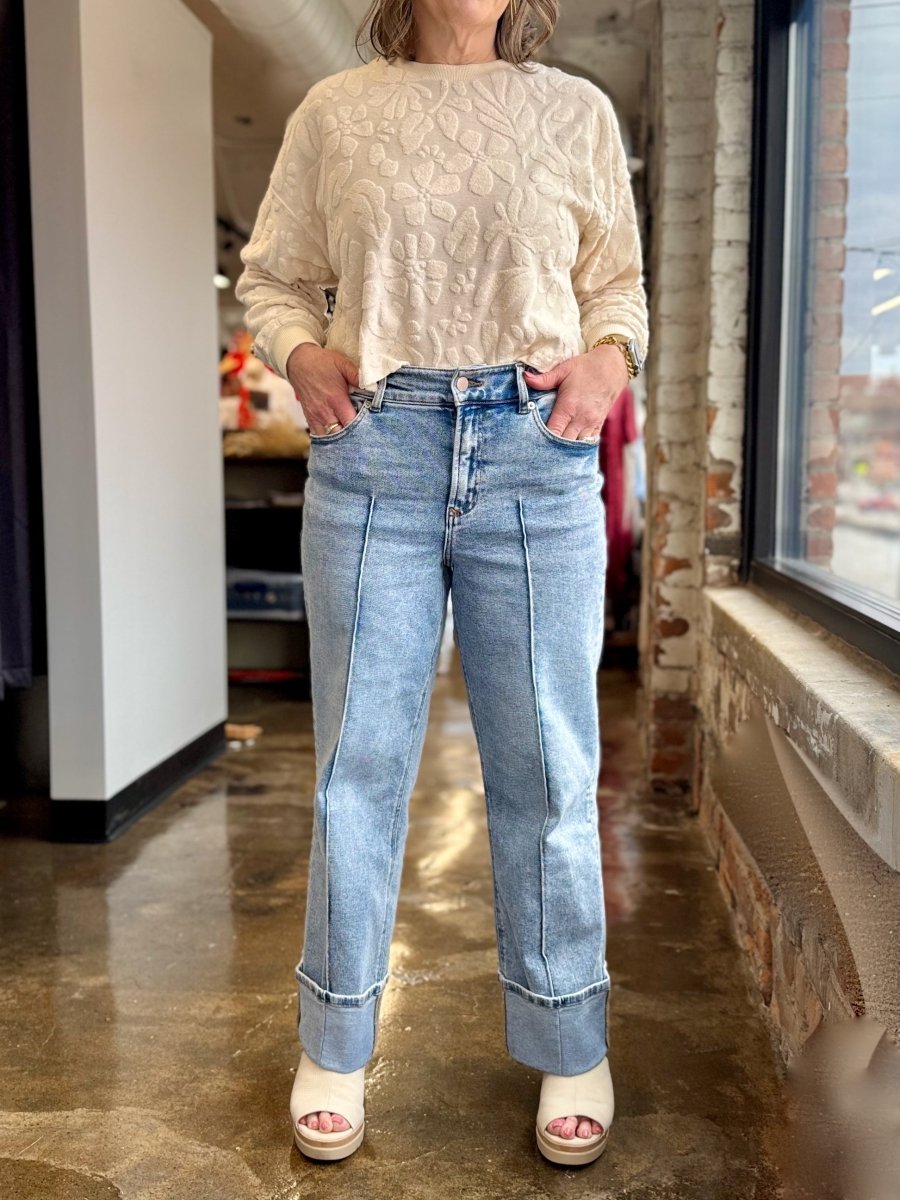 A person standing indoors wearing a cream textured long-sleeve top, light blue straight-leg jeans with rolled cuffs, and white platform sandals.