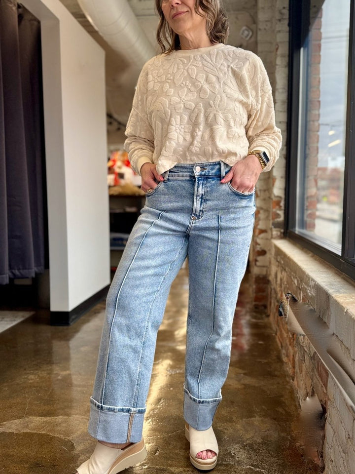 A woman wearing a cream cable-knit sweater and light-wash wide-leg blue jeans with rolled cuffs, standing in a retail store corridor.
