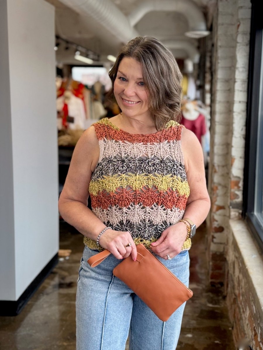 A woman wearing a sleeveless, multicolor knit top with horizontal stripes of rust orange, beige, gray, and yellow, paired with light blue jeans and holding a brown leather clutch in a store corridor.